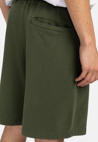 Green cotton shorts feature a relaxed fit, elastic waistband, and a rear patch pocket. Smooth texture with stitching details.