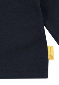 Maglietta a maniche lunghe in cotone blu navy con una texture liscia. Presenta un'etichetta gialla con lettering rosso all'orlo. Design semplice e minimalista.