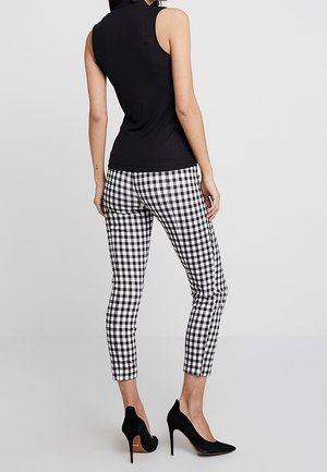 Nainen pukeutunut mustaan hihattomaan ribbineulepaitaan, mustavalkoisiin ruutukuviollisiin kapeisiin housuihin ja mustiin korkokenkiin, seisomassa yksivärisen taustan edessä.