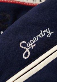 Tejido azul marino con texto bordado en blanco "Superdry", con acentos en color crema y una textura suave.