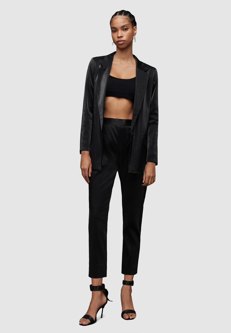 Schwarze Satinblazer über einem schwarzen Croptop, kombiniert mit hochgeschnittenen schwarzen Hosen und hochhackigen Sandalen mit Knöchelriemen.