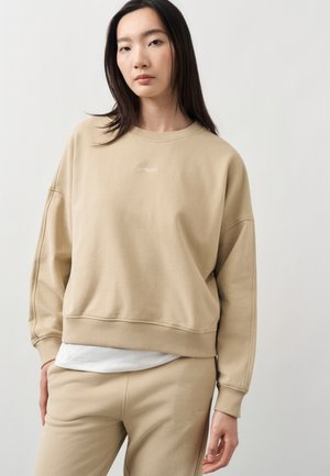 Tânără purtând un hanorac și pantaloni bej, cu o cămașă albă dedesubt, stând pe un fundal simplu, deschis.