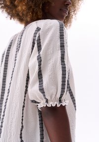 Witte getextureerde blouse met zwarte verticale strepen en geplooide mouwen met elastische manchetten. De stof heeft een subtiel geweven patroon.