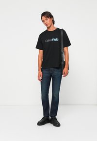 Čierne bavlnené tričko s logom "Calvin Klein" v modrej farbe, skombinované s tmavomodrými džínami a čiernymi topánkami. Model stojí na bielom pozadí.