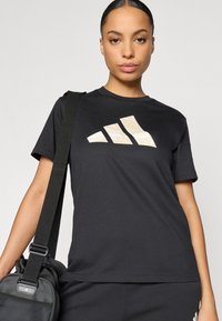 Černé bavlněné tričko s krátkými rukávy, na přední straně s velkým béžovým logem se třemi pruhy, doplněné černou taškou.