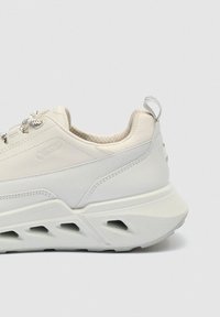 Svetlo krémová teniska s hrubou, tvarovanou podrážkou, golierom vyplneným medovitou štruktúrou, pútkom na päte a vzorovanými šnúrkami.