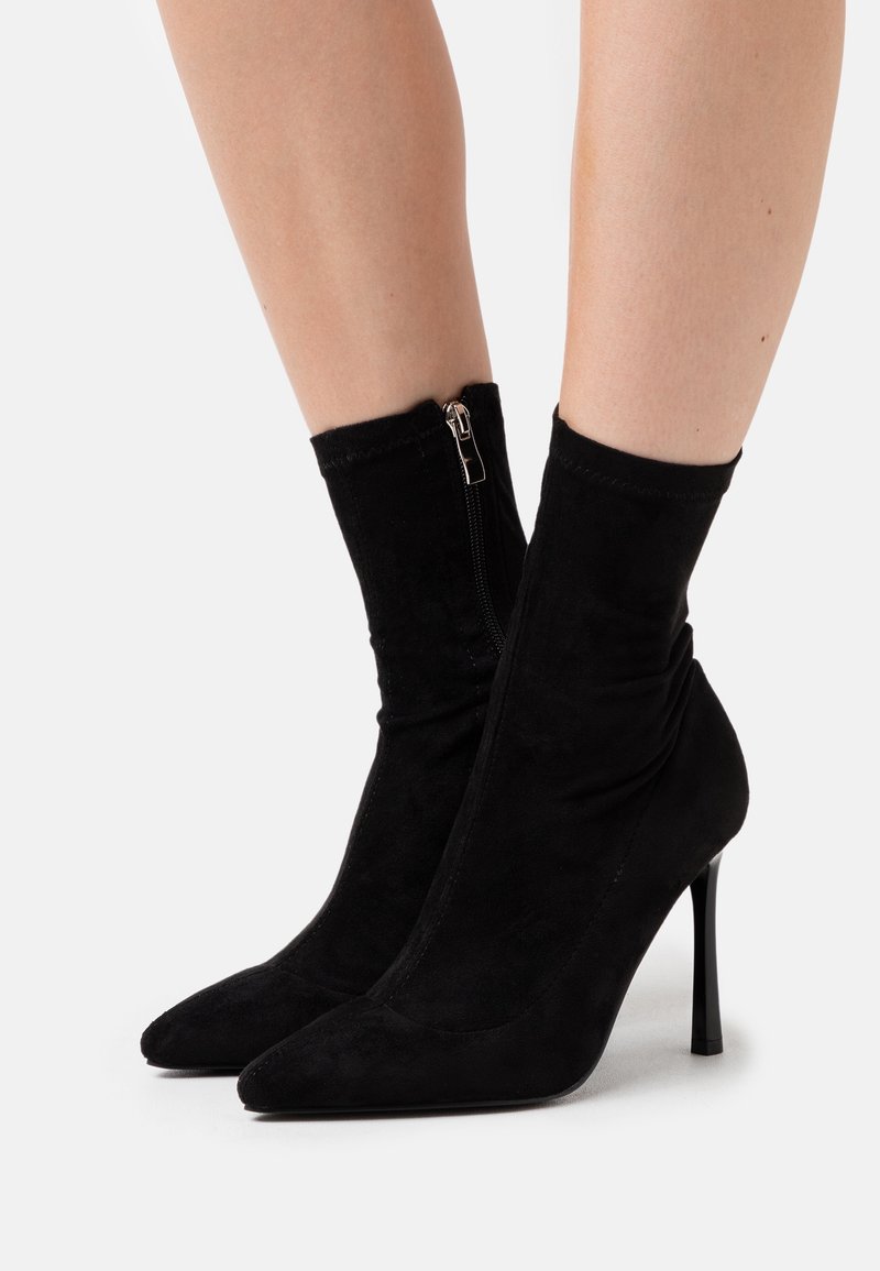 Bottines en daim noir avec bouts pointus, fermetures éclair latérales et talons stilettos portées sur des jambes nues sur un fond clair uni.