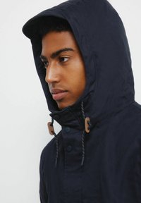 Giacca blu navy con cappuccio e cordoncino, caratterizzata da una texture liscia, due dettagli in legno marrone e chiusura con bottoni a pressione.