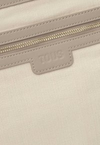 Beige textured fabric with herringbone pattern. Features a zippered closure and a prominent embossed "TOUS" logo.