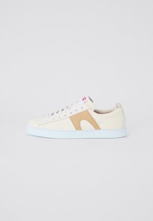 Low-top sneaker with a cream upper, tan accent, white sole, and lace-up design. Features a rear pull tab and zigzag stitching detail.