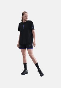 Kvinde står i en sort atletiktrøje og shorts med lilla detaljer, sorte Nike-sokker og sorte sneakers mod en hvid baggrund.