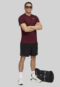 Camiseta roja de Puma, pantalones cortos negros, bolsa deportiva negra, calcetines blancos y zapatillas deportivas beige. Diseño elegante con la marca Puma.