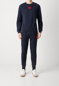 Set in felpa navy composto da felpa e pantaloni jogger, con toppe rosse su entrambi i capi, polsini a costine e design aderente. Abbinato a sneakers.
