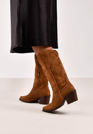 Botas de ante marrón hasta la rodilla con un ligero tacón, que presentan un intrincado bordado y un cierre de cremallera en el lateral. Suela lisa y puntera redonda.