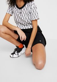 Person kniet in einem schwarz-weiß gemusterten Adidas-Shirt, schwarzen Shorts, orangenen Socken und schwarz-weißen Sneakern.