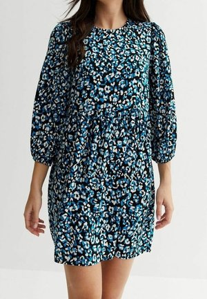 Femme portant une mini-robe ample à manches longues avec un motif léopard abstrait bleu, noir et blanc.