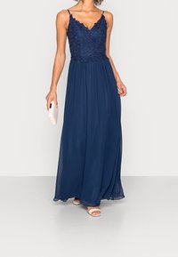 Žena oblečená v námořnickém modrém bezrukávovém krajkovém šatech, držící bílou kabelku a obutá v bílých sandálech na otevřenou špičku na jednobarevném pozadí.