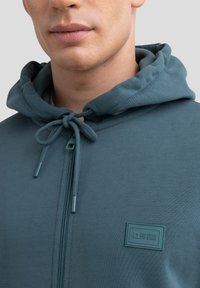 Felpa con zip color teal, dotata di cappuccio regolabile, caratterizzata da un tessuto testurizzato e una patch logo rettangolare. Finitura liscia con dettagli di cucitura sottili.