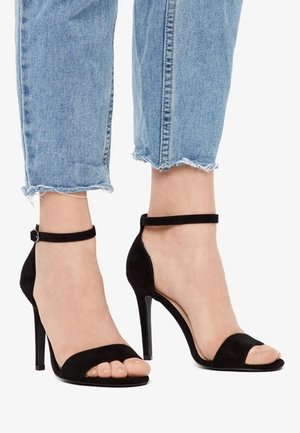 Person iført cropped blå jeans og sorte højhælede sandaler med ankelremme, stående på hvid baggrund.