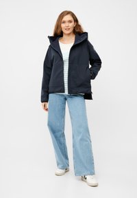 Giacca softshell blu navy con cappuccio, dotata di zip frontale, tasche laterali e vestibilità comoda. Indossata sopra una maglietta a righe e jeans.