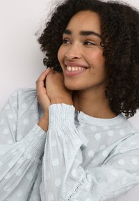 Lichtblauwe blouse met een gestructureerd bloemenpatroon, smockmanshouders en een knoopsluiting. De stof oogt zacht en lichtgewicht.