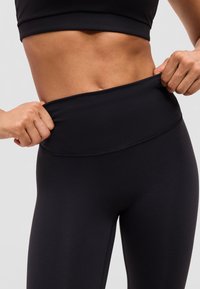 Wysportowana talia z rękami poprawiającymi czarne legginsy z wysokim stanem i nosząca czarny sportowy stanik na jednolitym tle.