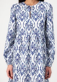 Rochie cu mânecă lungă din material alb, cu modele florale complicate în nuanțe de albastru și albastru deschis, cu decolteu în formă de ochi de fereastră și talie strânsă.