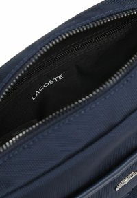 Borsa con cerniera di colore blu navy e trama, aperta per mostrare la fodera interna nera con il nome del marchio Lacoste stampato all'interno.
