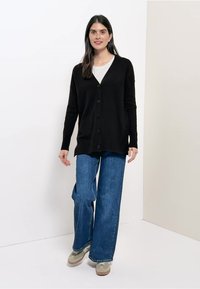 Zwarte gebreide cardigan met V-hals en knoopsluiting aan de voorkant, gedragen over een wit topje, gecombineerd met blauwe denim wijde broekspijpen en beige schoeisel.