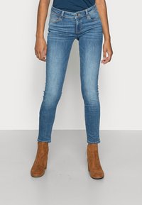 Jeans ajustados azul hechos de mezclilla, con un diseño clásico de cinco bolsillos, cierre frontal con botón y detalles de desvanecimiento sutil. Botines marrones. 