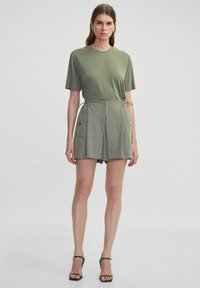Olivgrünes Baumwoll-T-Shirt mit kurzen Ärmeln, kombiniert mit hochgeschnittenen Shorts mit Taschen an den Seiten und Bindebändern, getragen mit schwarzen Riemchensandalen.