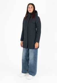Donkerblauwe parka met een capuchon, voorzien van een volledige ritssluiting, twee siervakken aan de voorkant en details van elastische boorden. Gekoeld met wijde spijkerbroeken en sneakers.