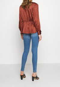 Blusa de estampado animal rojo con cintura de peplum, mangas largas y puños de botón, combinada con vaqueros ajustados azules y sandalias negras de tacón.