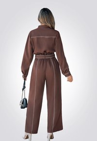 Mono marrón de manga larga, con cintura elástica y costuras blancas contrastantes. La modelo sostiene un bolso negro y lleva unos tacones nude.