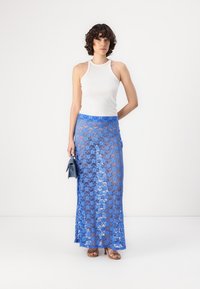 Jupe maxi en dentelle bleue transparente, avec un motif floral. Associée à un débardeur blanc et accessoirisée avec un petit sac à main bleu.