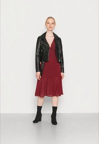 Jaqueta de motociclista cropped em couro preto sobre um vestido vermelho-escuro com bainha godê. Modelo usa botas de tornozelo pretas. Tecidos suaves, design discreto.