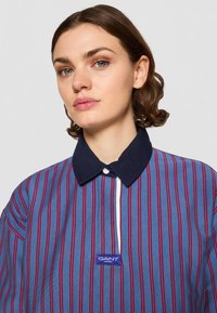 Camisa riscada verticalmente em azul e vermelho, com gola navy, apresentando um fecho por botões e uma etiqueta de logotipo azul na parte da frente.