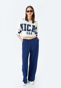 Hvid cropped sweatshirt med "CHICA U.S.A" i marineblå, parret med marineblå brede bukser. Modellen bærer smalle, mørke solbriller og hvide sko.