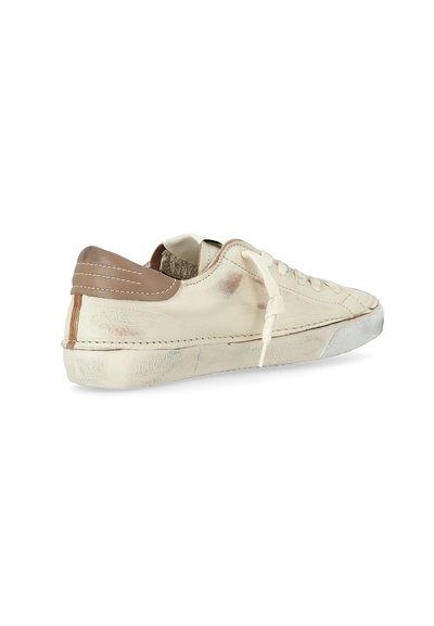 Sneaker in pelle bianca sporca con linguetta del tallone beige, chiusura con lacci e texture usurata, leggermente vissuta su uno sfondo bianco.