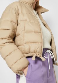 Beige puffjacka med en kortad passform, med horisontella kviltade sektioner, en framficka och dragskoekant; bärs över en grå tröja och lila byxor.