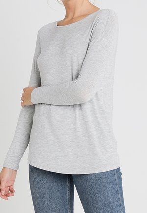 Kvinde iført en lysegrå langærmet skjorte og blå jeans, med den ene hånd hvilende på den modsatte arm mod en ensfarvet baggrund.