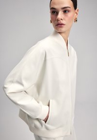 Giacca bomber bianca con colletto e polsini a coste, chiusura con zip e tasche laterali. Tessuto liscio e vestibilità rilassata con cuciture minime.