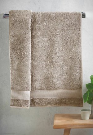 Asciugamano beige appeso a un porta asciugamani cromato sopra una panca in legno, accanto a una pianta verde in un vaso chiaro su uno sfondo di muro grigio.