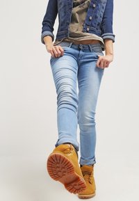 Světle modré úzké džíny z denimu, kombinované se šedou košilí a modrou džískou. Na nohou jsou hnědé šněrovací boty s texturovanou podrážkou.