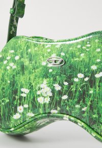 Sac à main vert avec un design floral présentant des marguerites blanches sur de l'herbe. Matériau synthétique lisse, forme arrondie et accent de logo en métal.