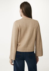 Beige stickad tröja med ribbat avslut och vida ärmar, med en mjuk textur. Kombinationen med mörkblå jeans är synlig bakifrån.