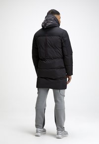 Giacca a vento nera imbottita con cappuccio, caratterizzata da un design trapuntato orizzontale, una tasca con zip sulla schiena e dettagli rossi a contrasto. Pantaloni grigi visibili.