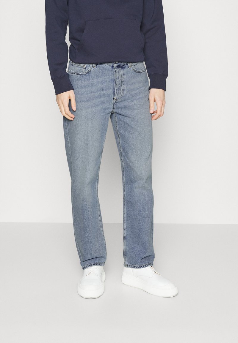 Personne portant un jean bleu clair droit, des baskets blanches et un sweat-shirt bleu foncé, debout devant un fond uni.