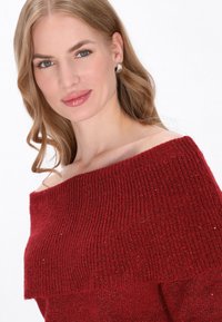 Pull rouge à épaules dénudées avec texture côtelée et détail de col replié. Tissu tricoté doux avec des accents de paillettes discrètes.
