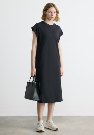 Νεαρή γυναίκα με μαύρο midi φόρεμα χωρίς μανίκια και λευκά αθλητικά παπούτσια, κρατώντας μια μαύρη τσάντα, στέκεται μπροστά σε ένα απλό γκρίζο φόντο.